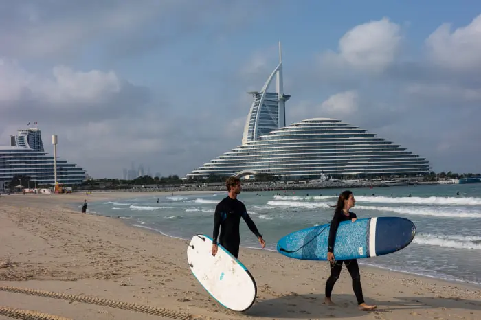Two people in wetsuits carry surfboards on Jumeirah Beach with the Burj al Arab hotel in the background.